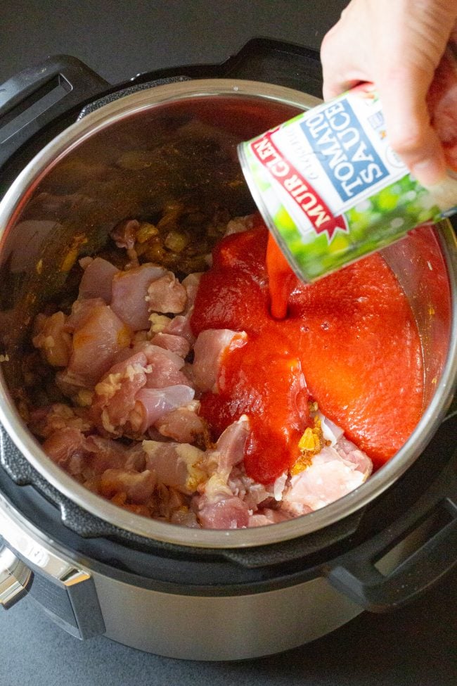 Raw chicken in an instant pot with a hand pouring in tomato sauce. 